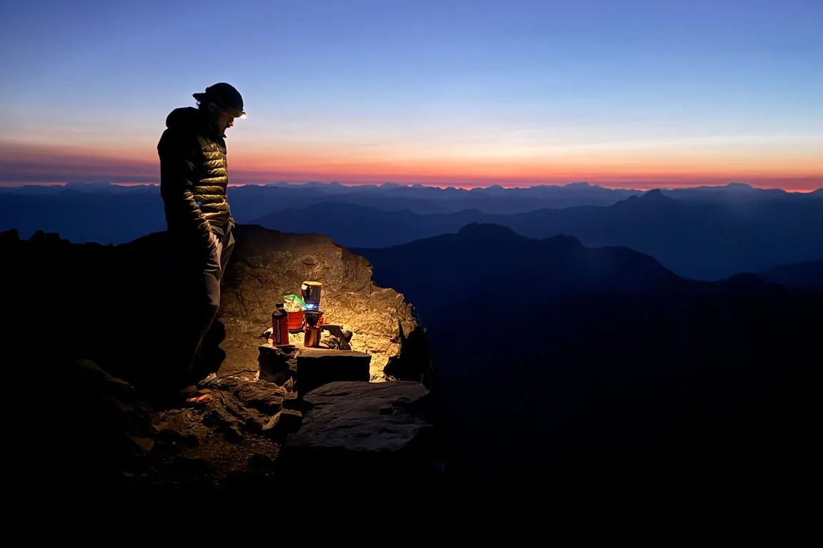 A camper beside a stove and cooking supplies as sunrise lights the mountain horizon.