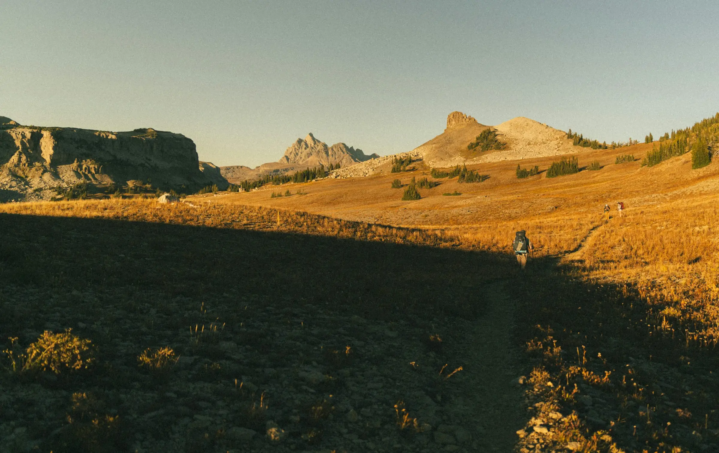 A trail crosses golden mountain grass at dusk, with small hikers in the distance.